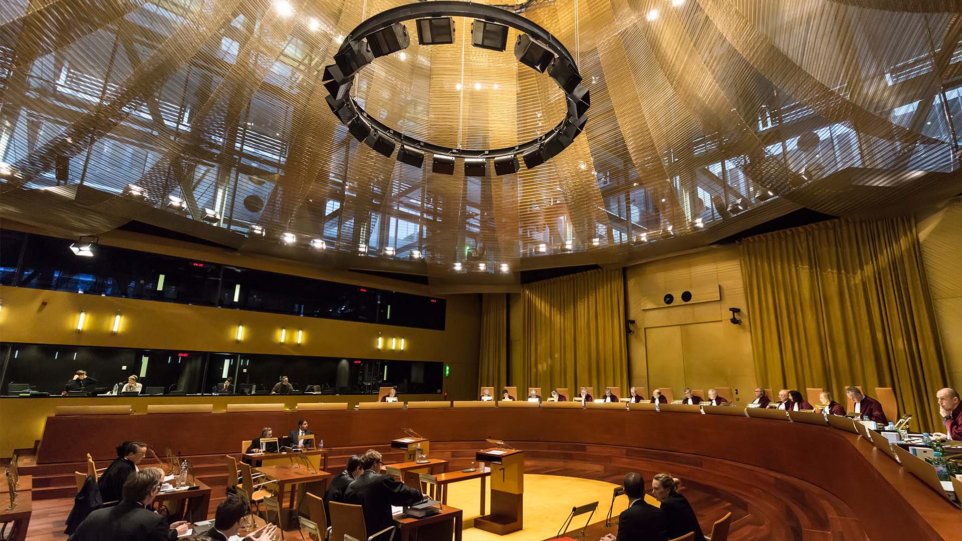 Audiencia en la Gran Sala del Tribunal de Justicia de la Unión Europea, con jueces sentados en un estrado curvo y abogados en el área central, bajo una gran estructura circular de iluminación. Inteligencia artificial