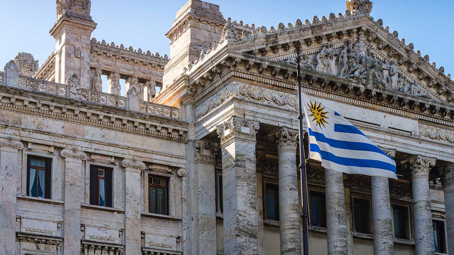 Fachada del Palacio Legislativo de Montevideo, un edificio de estilo neoclásico ubicado en Montevideo, Uruguay, que alberga el Parlamento del país. La imagen muestra una vista frontal del edificio, con una bandera del propio país ondeando delante. Elecciones en Uruguay