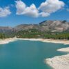 Imagen diurna del embalse de Guadalest (Alicante). En primer plano, el agua del embalse y la orilla. Al fondo, un bosque con árboles verdes y las montañas bajo el cielo azul con algunas nubes blancas. Agua en Europa