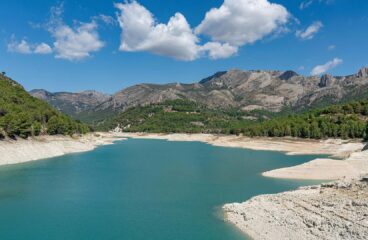 Imagen diurna del embalse de Guadalest (Alicante). En primer plano, el agua del embalse y la orilla. Al fondo, un bosque con árboles verdes y las montañas bajo el cielo azul con algunas nubes blancas. Agua en Europa