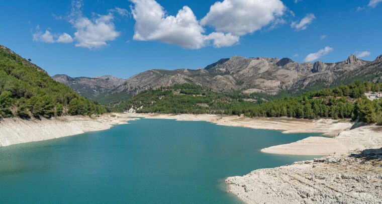 Imagen diurna del embalse de Guadalest (Alicante). En primer plano, el agua del embalse y la orilla. Al fondo, un bosque con árboles verdes y las montañas bajo el cielo azul con algunas nubes blancas. Agua en Europa