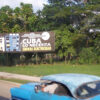 Cartel en una calle de Cuba con el mensaje "Cuba lo necesita, ahorra electricidad", mostrando edificios y un fondo de árboles. En primer plano, se observa un coche la parte superior de un coche azul. Cuba