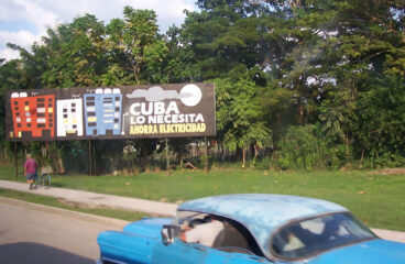 Cartel en una calle de Cuba con el mensaje "Cuba lo necesita, ahorra electricidad", mostrando edificios y un fondo de árboles. En primer plano, se observa un coche la parte superior de un coche azul. Cuba