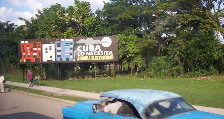 Cartel en una calle de Cuba con el mensaje "Cuba lo necesita, ahorra electricidad", mostrando edificios y un fondo de árboles. En primer plano, se observa un coche la parte superior de un coche azul. Cuba