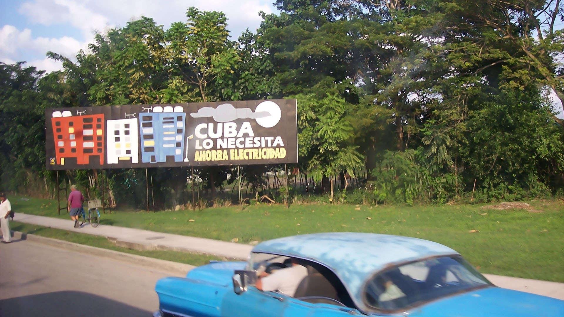 Cartel en una calle de Cuba con el mensaje "Cuba lo necesita, ahorra electricidad", mostrando edificios y un fondo de árboles. En primer plano, se observa un coche la parte superior de un coche azul. Cuba