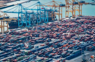 Vista aérea diurna de la zona industrial del puerto de Barcelona. En primer plano, se aprecian contenedores de varios colores y varias grúas para transportarlas. En el fondo, el mar Mediterráneo bajo el cielo azul. Pymes