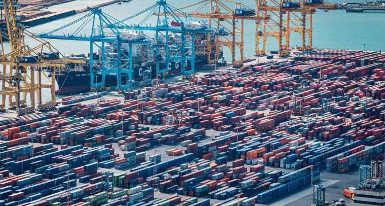 Vista aérea diurna de la zona industrial del puerto de Barcelona. En primer plano, se aprecian contenedores de varios colores y varias grúas para transportarlas. En el fondo, el mar Mediterráneo bajo el cielo azul. Pymes