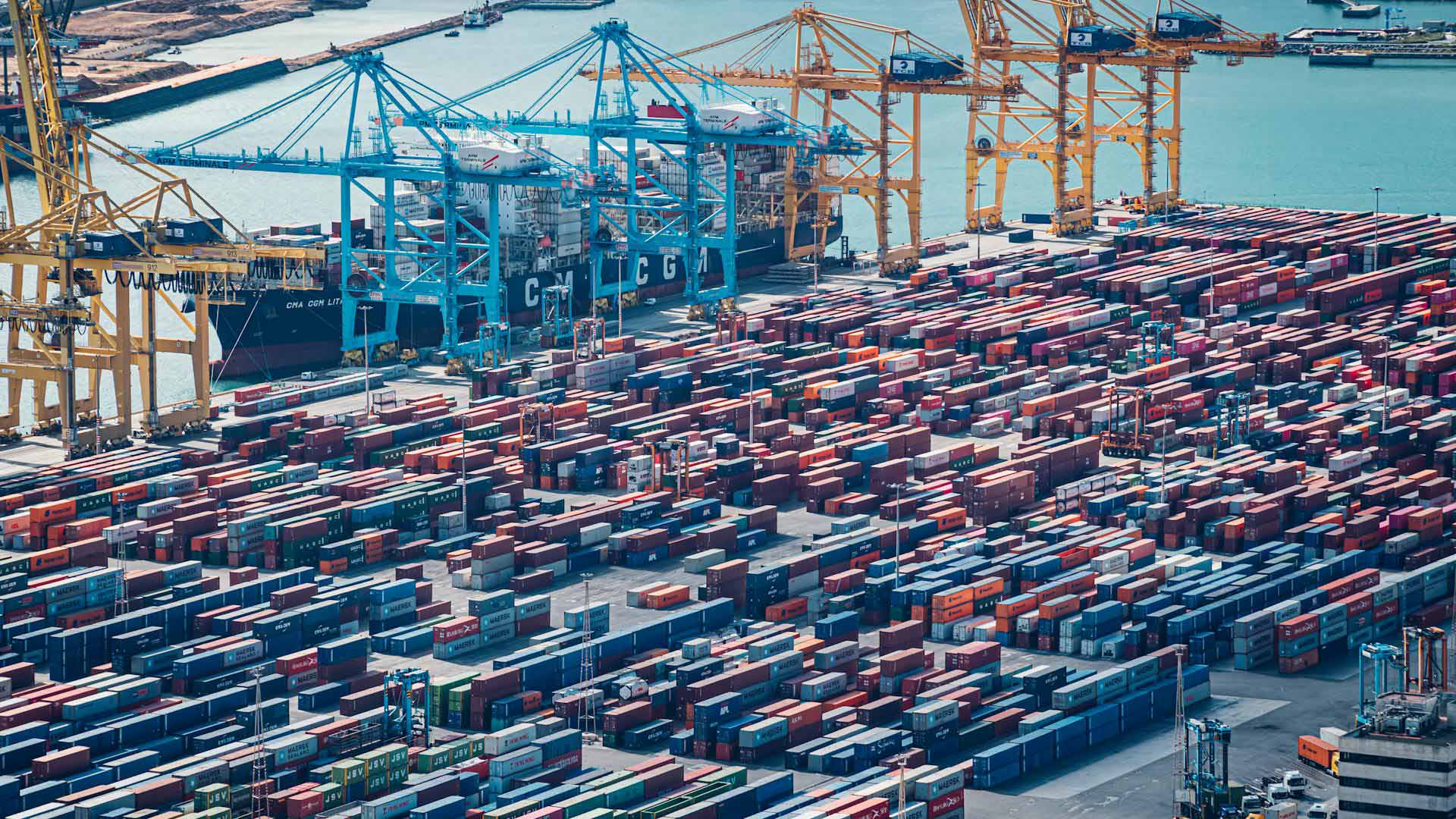 Vista aérea diurna de la zona industrial del puerto de Barcelona. En primer plano, se aprecian contenedores de varios colores y varias grúas para transportarlas. En el fondo, el mar Mediterráneo bajo el cielo azul. Pymes