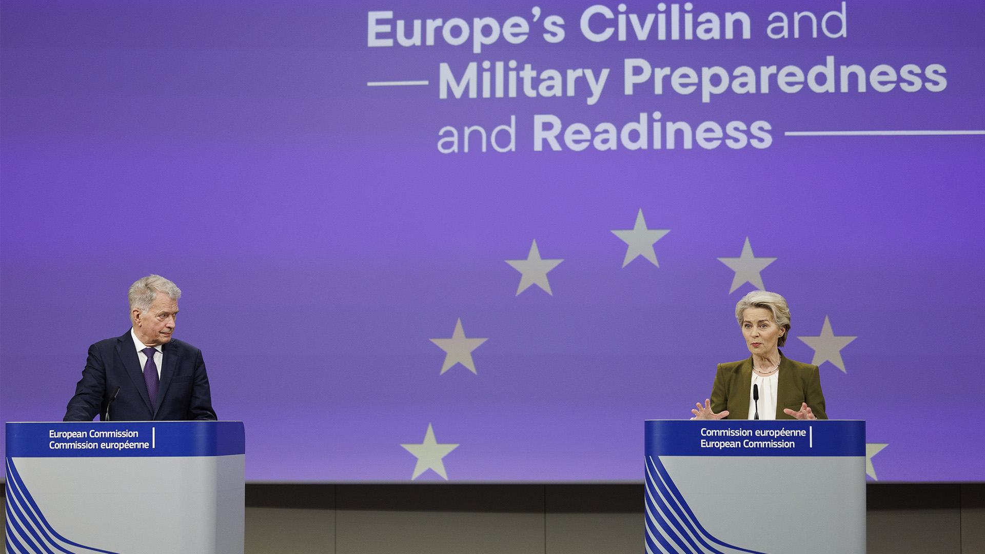 El expresidente de Finlandia, Sauli Niinistö, a la izquierda, y la presidenta de la Comisión Europea, Ursula von der Leyen, a la derecha en la rueda de prensa de presentación del Informe Niinistö. Ambos se encuentra de pie en podios frente a un fondo azul con estrellas amarillas que representan la bandera de la UE y el texto en inglés 'Strengthening Europe’s Civilian and Military Preparedness and Readiness', ‘’Refuerzo de la preparación civil y militar de Europa’’, en español. Niinistö