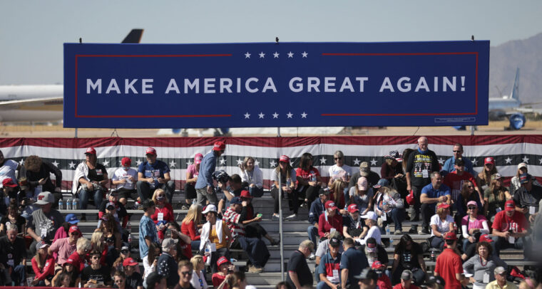 Seguidores de Donald Trump en un mitin diurno de campaña 'Make America Great Again' en el aeropuerto de Phoenix Goodyear, Arizona (EEUU). En primer plano, un cartel azul con el lema 'Make America Great Again' (Hagamos a América grande de nuevo) en letras blancas; al fondo, partes de aviones. Victoria de Trump