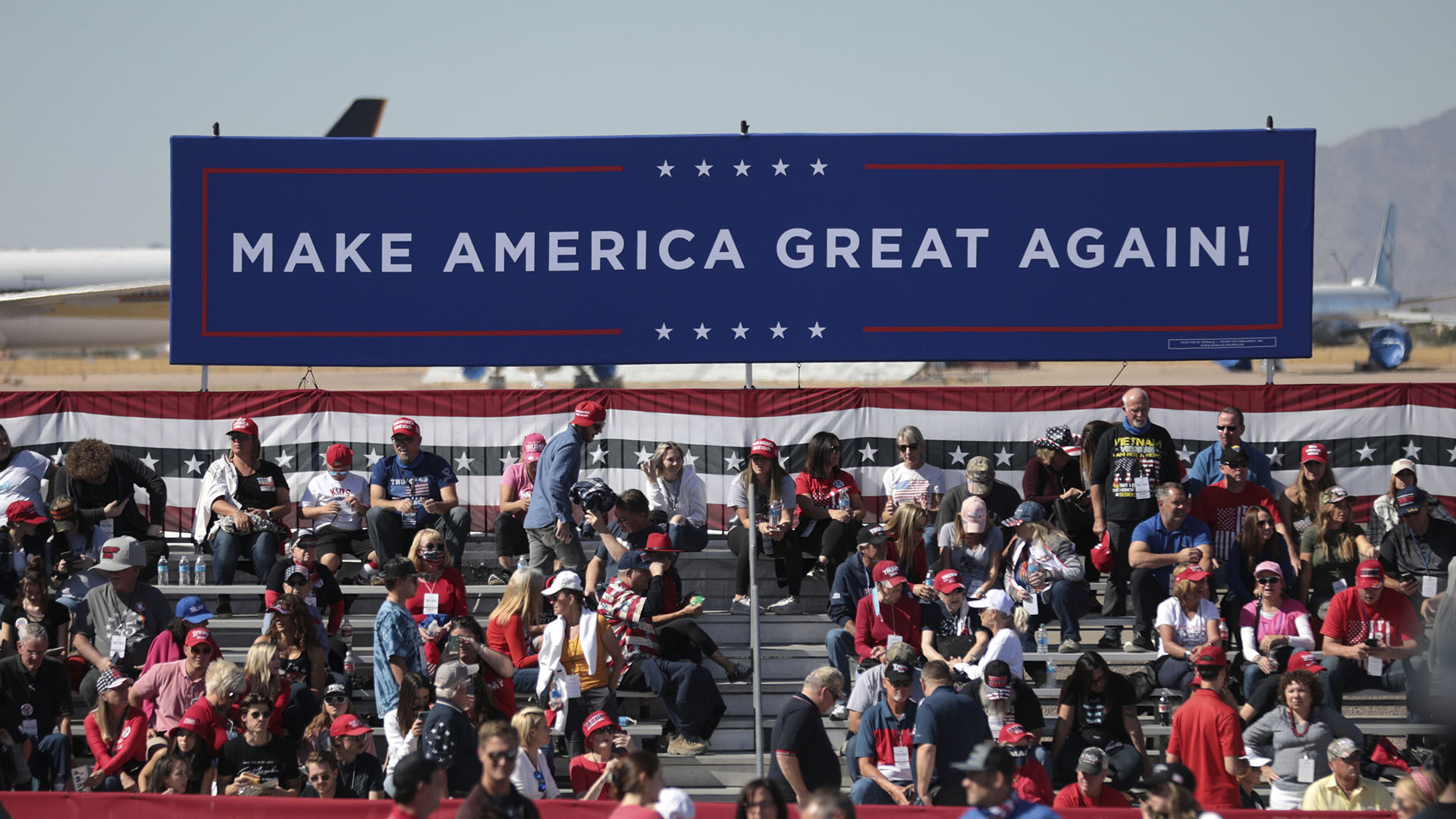 Seguidores de Donald Trump en un mitin diurno de campaña 'Make America Great Again' en el aeropuerto de Phoenix Goodyear, Arizona (EEUU). En primer plano, un cartel azul con el lema 'Make America Great Again' (Hagamos a América grande de nuevo) en letras blancas; al fondo, partes de aviones. Victoria de Trump