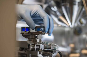 Mano de un técnico con guantes azules ajustando un componente en un equipo de laboratorio de alta precisión, rodeado de tecnología avanzada. Competitividad