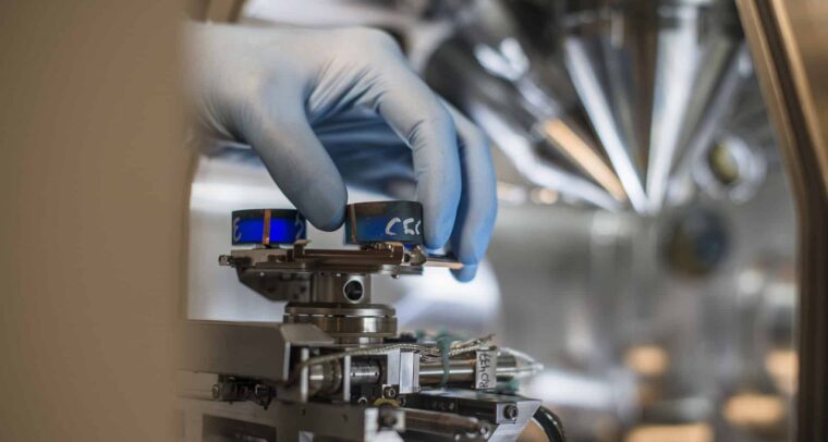 Mano de un técnico con guantes azules ajustando un componente en un equipo de laboratorio de alta precisión, rodeado de tecnología avanzada. Competitividad