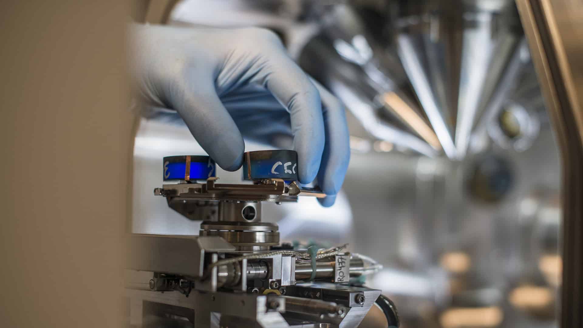 Mano de un técnico con guantes azules ajustando un componente en un equipo de laboratorio de alta precisión, rodeado de tecnología avanzada. Competitividad