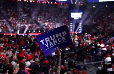 Cartel con el mensaje "Trump Vance: Make America Great Again!" sostenido por un asistente en un mitin político lleno de personas en un estadio iluminado. Trump