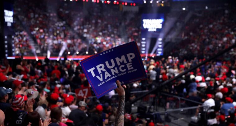 Cartel con el mensaje "Trump Vance: Make America Great Again!" sostenido por un asistente en un mitin político lleno de personas en un estadio iluminado. Trump