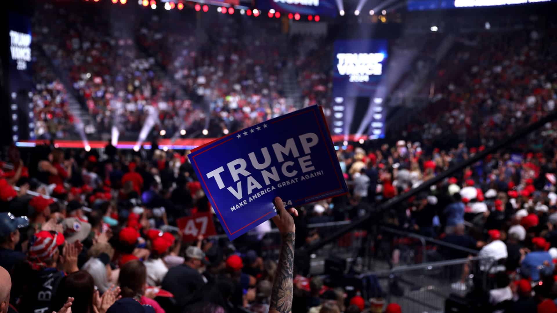 Cartel con el mensaje "Trump Vance: Make America Great Again!" sostenido por un asistente en un mitin político lleno de personas en un estadio iluminado. Trump