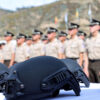 Cascos tácticos de color negro, colocados sobre una mesa blanca en primer plano, mientras que, en el fondo desenfocado, se observa a un grupo de oficiales de la Policía Nacional del Ecuador, uniformados y en formación, frente a un paisaje montañoso con banderas ondeando a lo lejos. Crimen organizado