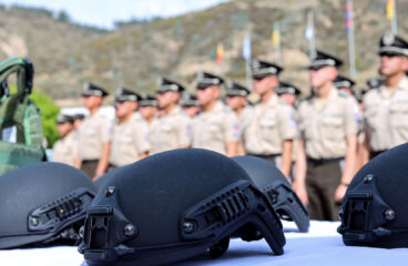 Cascos tácticos de color negro, colocados sobre una mesa blanca en primer plano, mientras que, en el fondo desenfocado, se observa a un grupo de oficiales de la Policía Nacional del Ecuador, uniformados y en formación, frente a un paisaje montañoso con banderas ondeando a lo lejos. Crimen organizado