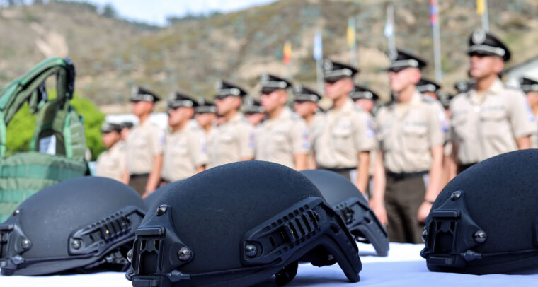 Cascos tácticos de color negro, colocados sobre una mesa blanca en primer plano, mientras que, en el fondo desenfocado, se observa a un grupo de oficiales de la Policía Nacional del Ecuador, uniformados y en formación, frente a un paisaje montañoso con banderas ondeando a lo lejos. Crimen organizado