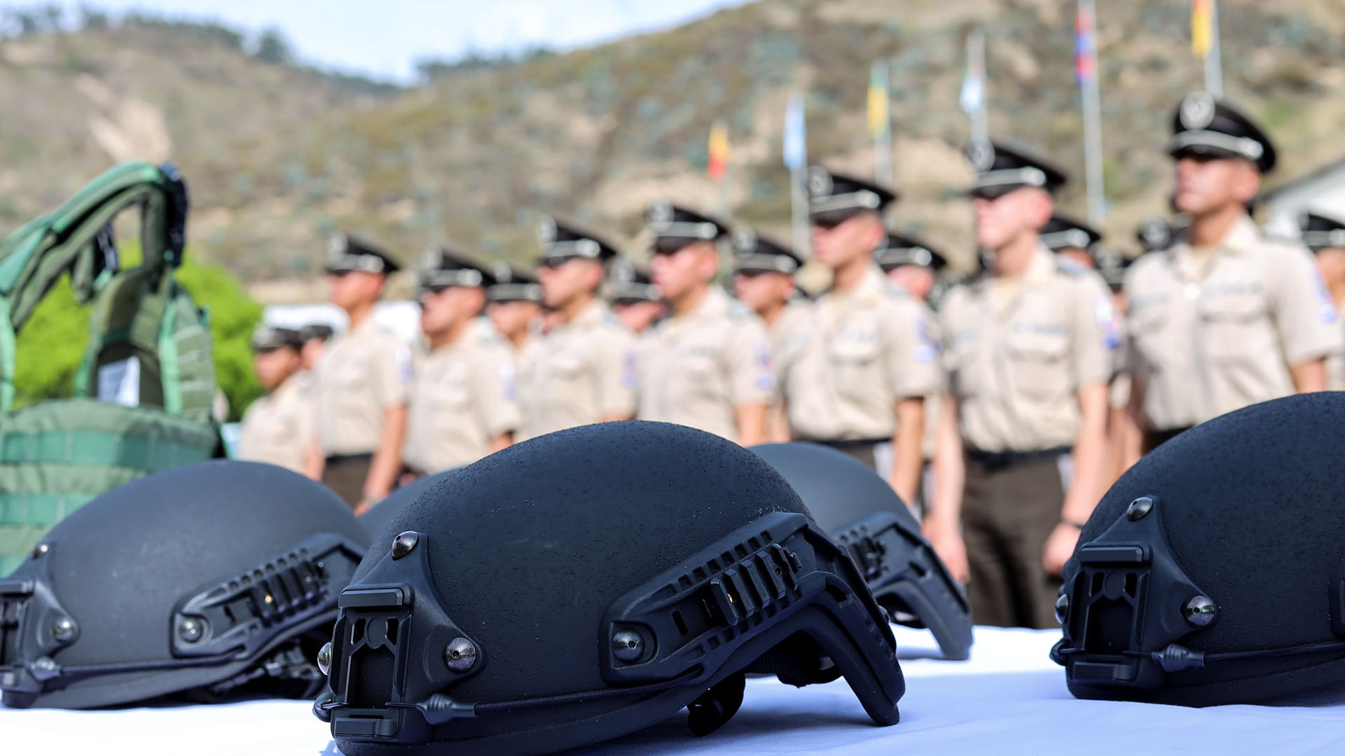 Cascos tácticos de color negro, colocados sobre una mesa blanca en primer plano, mientras que, en el fondo desenfocado, se observa a un grupo de oficiales de la Policía Nacional del Ecuador, uniformados y en formación, frente a un paisaje montañoso con banderas ondeando a lo lejos. Crimen organizado