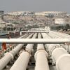 Vista general del puerto de la isla de Kharg con tuberías de petróleo en primer plano, instalaciones de almacenamiento y tanques etiquetados con "Persian Gulf" visibles al fondo, rodeados de un paisaje árido. irán