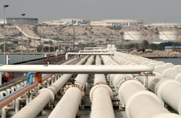 Vista general del puerto de la isla de Kharg con tuberías de petróleo en primer plano, instalaciones de almacenamiento y tanques etiquetados con "Persian Gulf" visibles al fondo, rodeados de un paisaje árido. irán