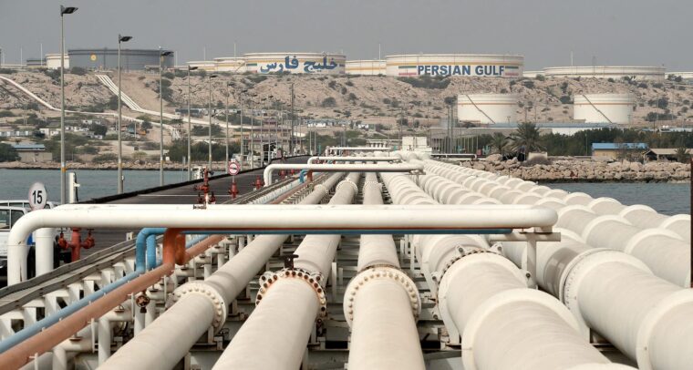 Vista general del puerto de la isla de Kharg con tuberías de petróleo en primer plano, instalaciones de almacenamiento y tanques etiquetados con "Persian Gulf" visibles al fondo, rodeados de un paisaje árido. irán