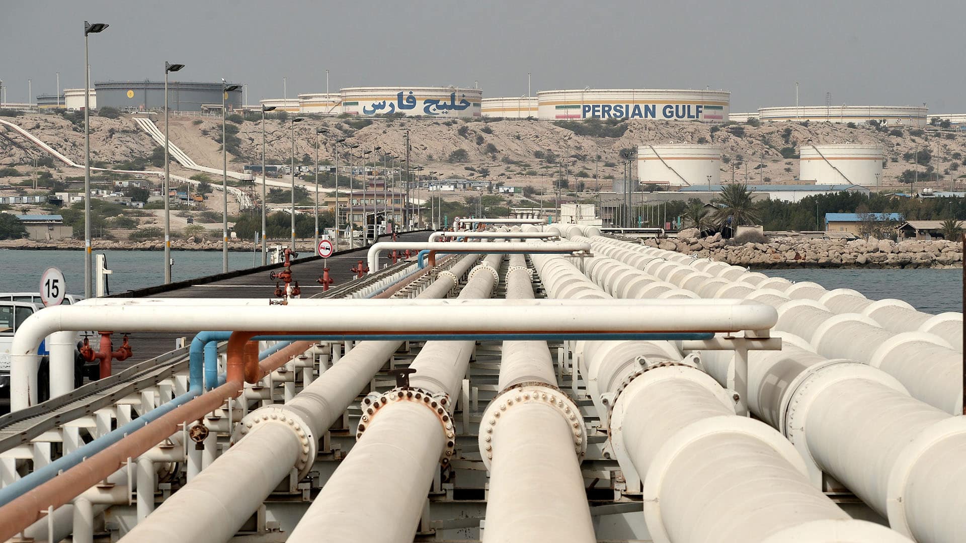 Vista general del puerto de la isla de Kharg con tuberías de petróleo en primer plano, instalaciones de almacenamiento y tanques etiquetados con "Persian Gulf" visibles al fondo, rodeados de un paisaje árido. irán