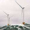 Offshore white wind turbines in rough ocean conditions, with large waves crashing against their bases under a cloudy sky. renewable energy