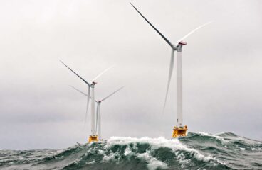 : Aerogeneradores blancos en alta mar en condiciones de océano agitado, con grandes olas chocando contra sus bases bajo un cielo nublado. Cambio climático