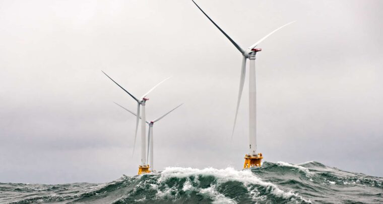 Offshore white wind turbines in rough ocean conditions, with large waves crashing against their bases under a cloudy sky. renewable energy