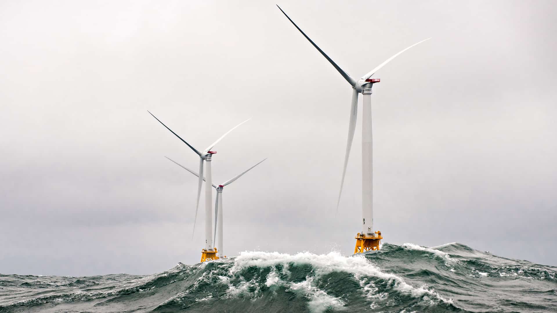 : Aerogeneradores blancos en alta mar en condiciones de océano agitado, con grandes olas chocando contra sus bases bajo un cielo nublado. Cambio climático