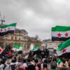 Multitud reunida en Trafalgar Square (Londres), con personas sosteniendo banderas con franjas verde, blanca y negra con tres estrellas rojas. En el fondo, se ven edificios históricos y una fuente bajo el cielo nublado. Bashar al-Assad