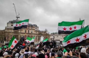 Multitud reunida en Trafalgar Square (Londres), con personas sosteniendo banderas con franjas verde, blanca y negra con tres estrellas rojas. En el fondo, se ven edificios históricos y una fuente bajo el cielo nublado. Bashar al-Assad