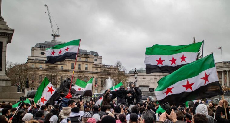 Multitud reunida en Trafalgar Square (Londres), con personas sosteniendo banderas con franjas verde, blanca y negra con tres estrellas rojas. En el fondo, se ven edificios históricos y una fuente bajo el cielo nublado. Bashar al-Assad