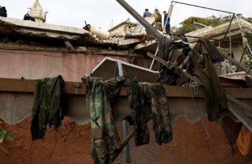 Primer plano de un edificio destruido en el Líbano, con escombros y ropa militar colgada. En el fondo, personas inspeccionan los datos sobre el techo a la luz de un día nublado. Líbano