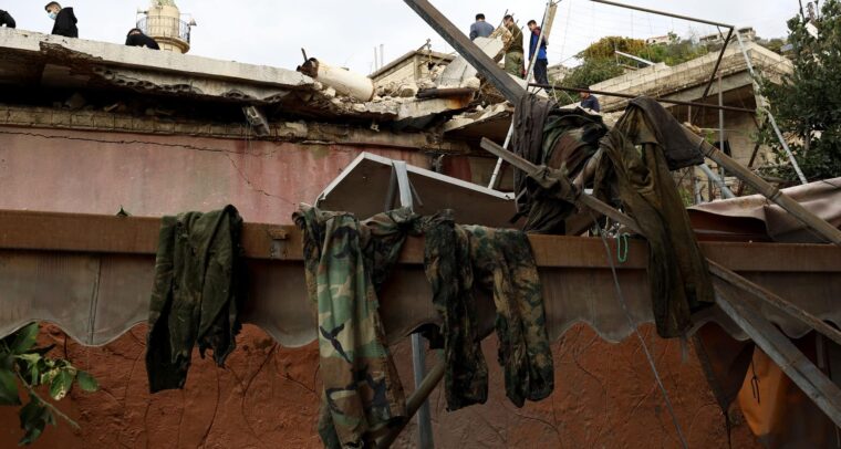 Primer plano de un edificio destruido en el Líbano, con escombros y ropa militar colgada. En el fondo, personas inspeccionan los datos sobre el techo a la luz de un día nublado. Líbano