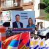 Vehículo decorado con carteles de campaña electoral del Frente Amplio, que muestra a Yamandú Orsi como candidato a presidente y Carolina Cosse como candidata a vicepresidenta. Delante del vehículo, se observan banderas de Uruguay y del Frente Amplio, junto a conos naranjas y transeúntes en una calle. Uruguay