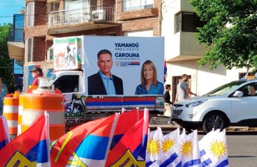 Vehículo decorado con carteles de campaña electoral del Frente Amplio, que muestra a Yamandú Orsi como candidato a presidente y Carolina Cosse como candidata a vicepresidenta. Delante del vehículo, se observan banderas de Uruguay y del Frente Amplio, junto a conos naranjas y transeúntes en una calle. Uruguay