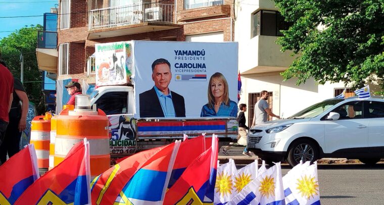 Vehículo decorado con carteles de campaña electoral del Frente Amplio, que muestra a Yamandú Orsi como candidato a presidente y Carolina Cosse como candidata a vicepresidenta. Delante del vehículo, se observan banderas de Uruguay y del Frente Amplio, junto a conos naranjas y transeúntes en una calle. Uruguay