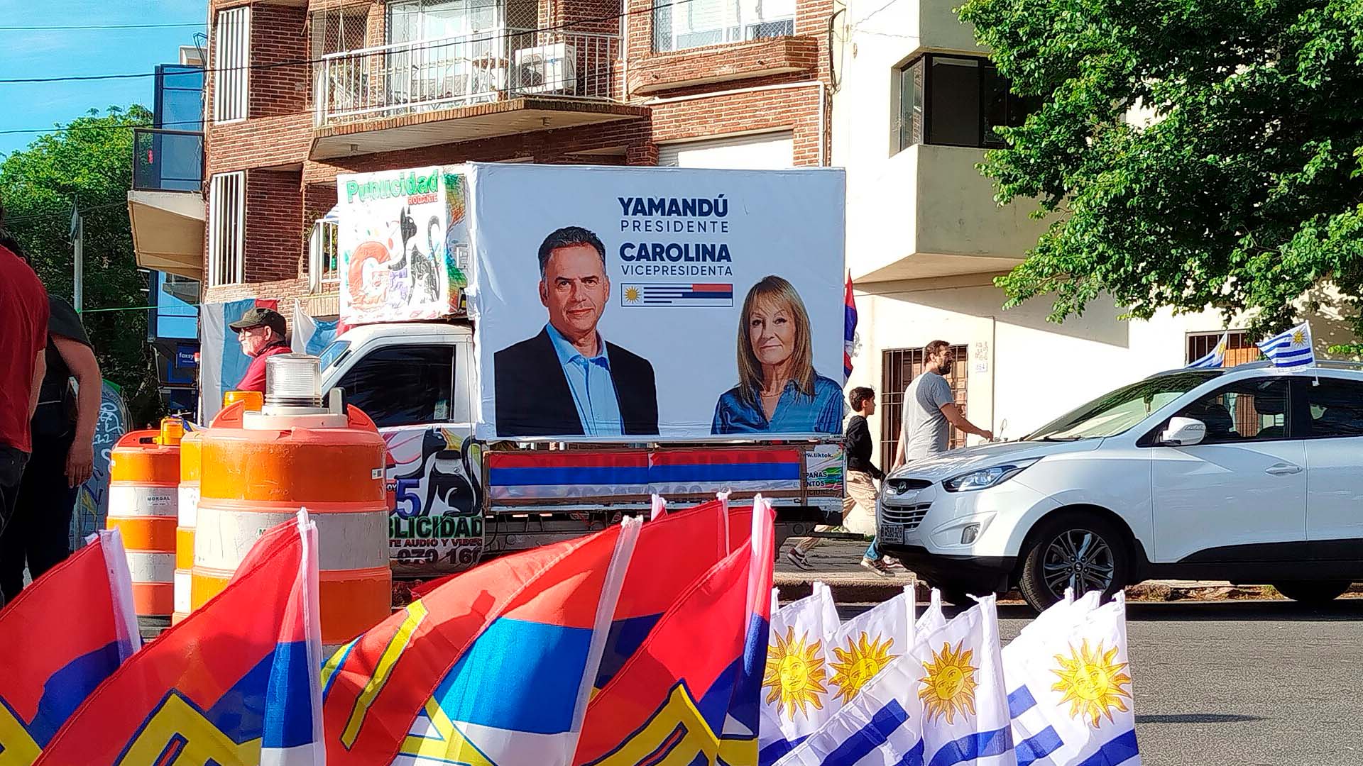 Vehículo decorado con carteles de campaña electoral del Frente Amplio, que muestra a Yamandú Orsi como candidato a presidente y Carolina Cosse como candidata a vicepresidenta. Delante del vehículo, se observan banderas de Uruguay y del Frente Amplio, junto a conos naranjas y transeúntes en una calle. Uruguay