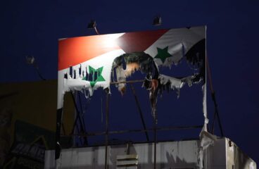 Cartel dañado de Bashar al-Assad con la bandera de Siria de fondo mostrando signos de deterioro y quemaduras, con un cielo nocturno al fondo