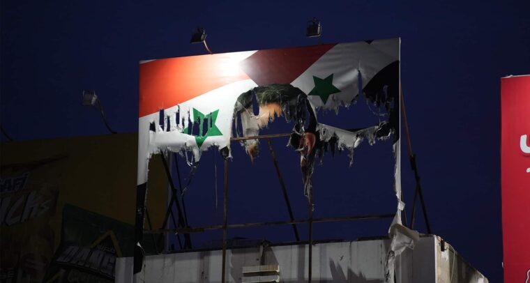 Cartel dañado de Bashar al-Assad con la bandera de Siria de fondo mostrando signos de deterioro y quemaduras, con un cielo nocturno al fondo