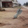 Una calle desierta con edificios dañados a ambos lados en la ciudad siria de Taftanaz. En el primer plano, se ve una parte de un proyectil de artillería incrustada en el suelo, con escombros y cables dispersos alrededor. Siria