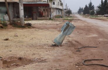 Una calle desierta con edificios dañados a ambos lados en la ciudad siria de Taftanaz. En el primer plano, se ve una parte de un proyectil de artillería incrustada en el suelo, con escombros y cables dispersos alrededor. Siria