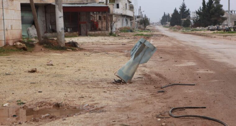 Una calle desierta con edificios dañados a ambos lados en la ciudad siria de Taftanaz. En el primer plano, se ve una parte de un proyectil de artillería incrustada en el suelo, con escombros y cables dispersos alrededor. Siria