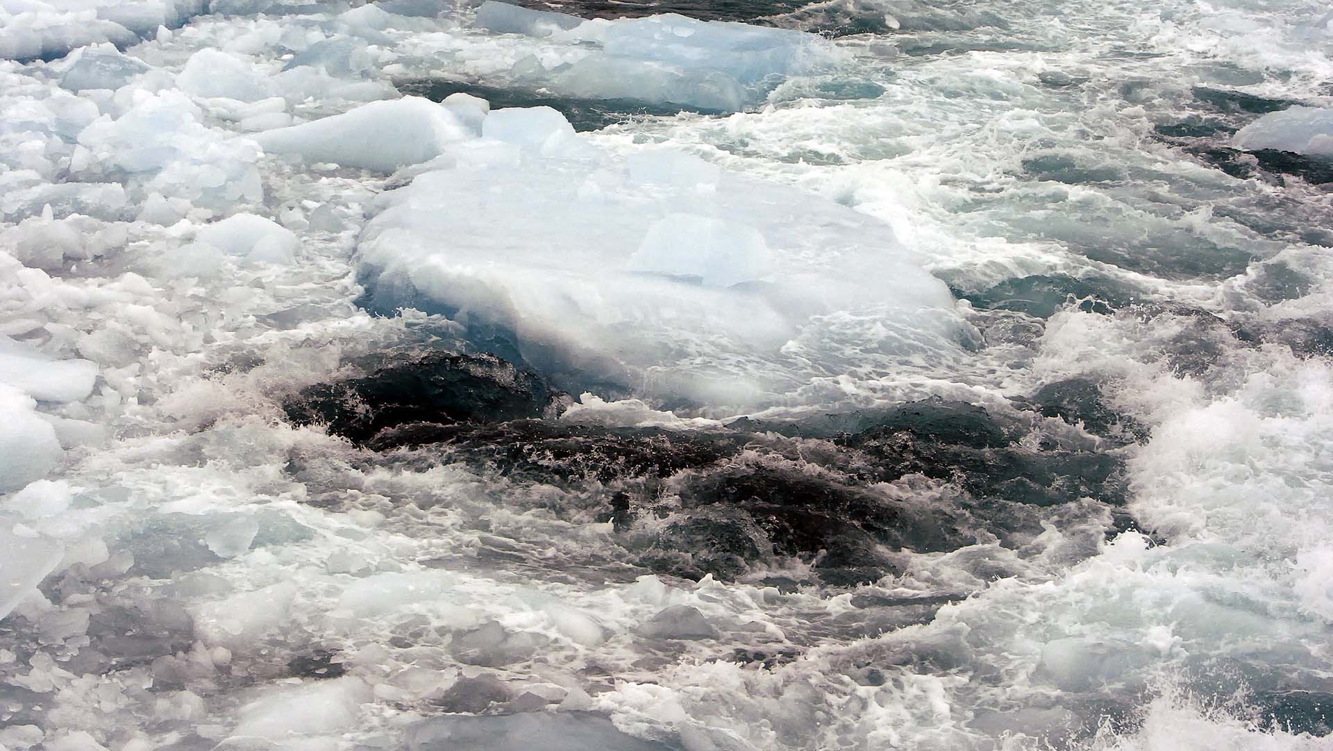 Hielo marino fragmentado flotando en aguas turbulentas y frías, mostrando una mezcla de bloques de hielo azules y espuma blanca debido a las corrientes marinas. Agua