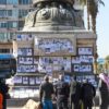 Personas reunidas alrededor de una estructura donde cuelgan fotografías de familiares desaparecidos, en una plaza urbana en Alepo (Siria). Se observan edificios, palmeras y varias personas mirando las imágenes y oraciones expuestas. HTS