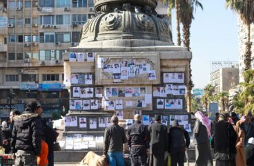 Personas reunidas alrededor de una estructura donde cuelgan fotografías de familiares desaparecidos, en una plaza urbana en Alepo (Siria). Se observan edificios, palmeras y varias personas mirando las imágenes y oraciones expuestas. HTS